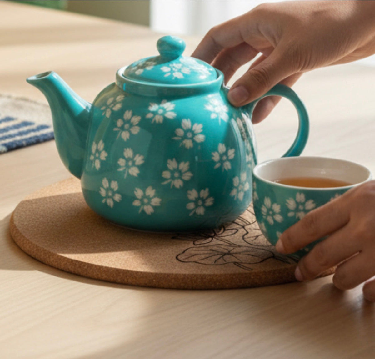 Turquoise teapot with white floral pattern and matching cup on a cork trivet.