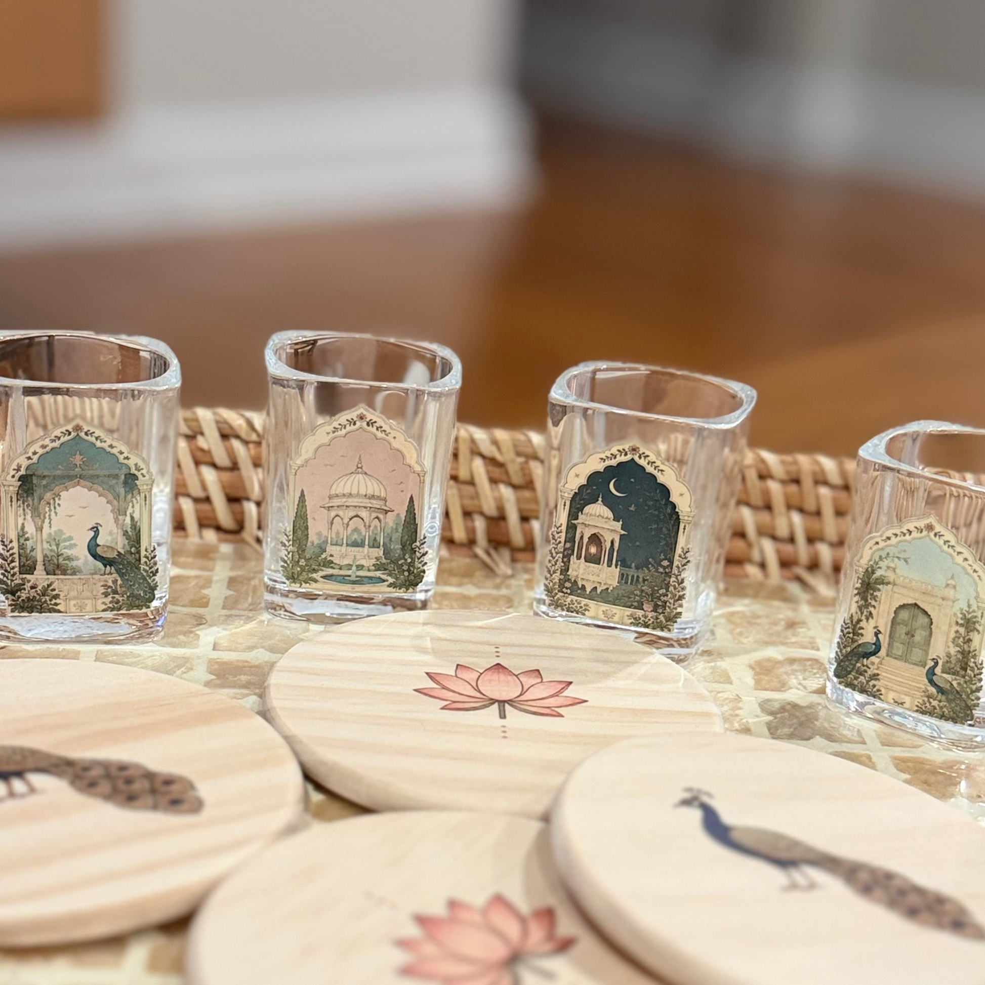 A set of decorative glass shot glasses and round wooden coasters with peacock and lotus illustrations, arranged in a woven wicker tray on a wooden table.